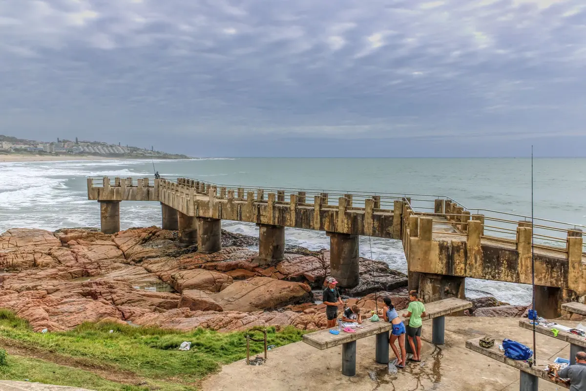 Famille multigénérationnelle sur une jetée côtière face à la mer, valises et sacs de plage à proximité.